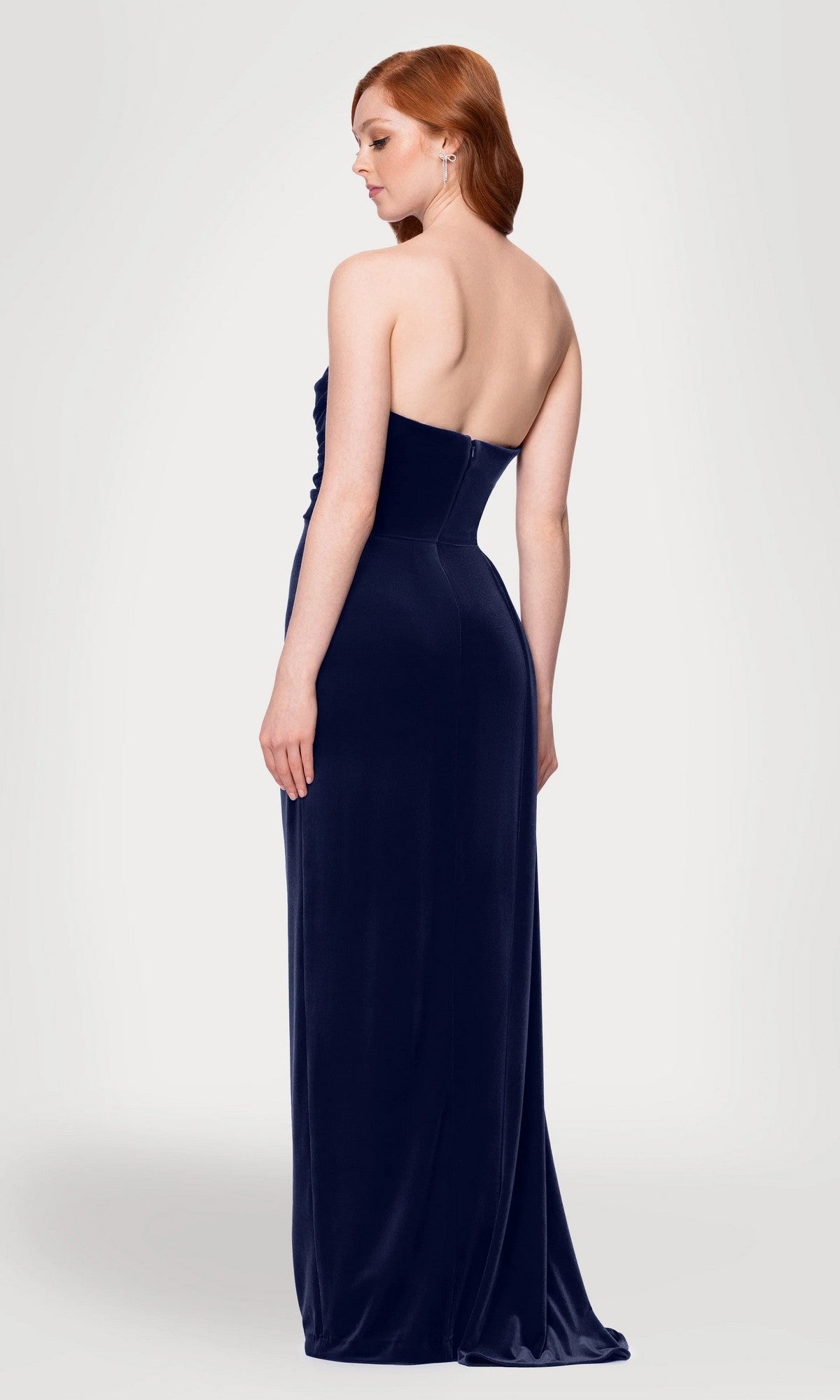 A woman with red hair stands with her back to the camera against a plain light background, wearing the Blondie Nites 5138BN long prom dress—a strapless velvet gown with a floor-length sash.